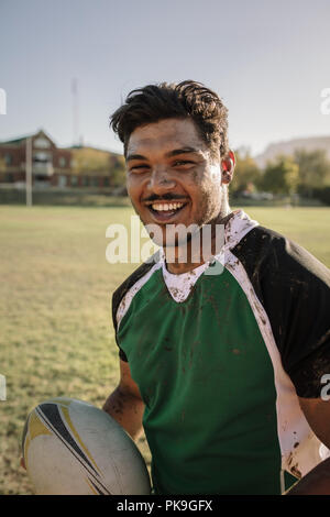 Happy young sportsman avec un ballon de rugby sur le terrain de sport. Smiling rugby player with ball traînées dans la boue. Banque D'Images