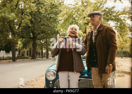 Smiling old couple debout devant leur voiture et parlant du paysage. Aîné gai l'homme et la femme dans des vêtements chauds en prenant des photographies Banque D'Images
