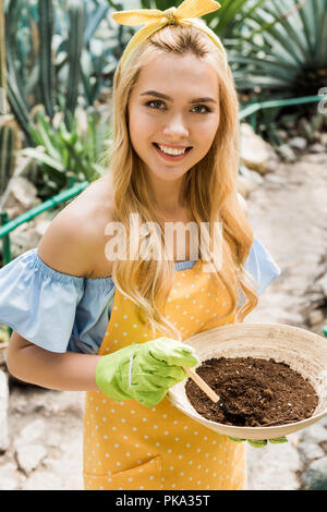 High angle view of beautiful blonde girl holding bowl avec sol et smiling at camera in greenhouse Banque D'Images