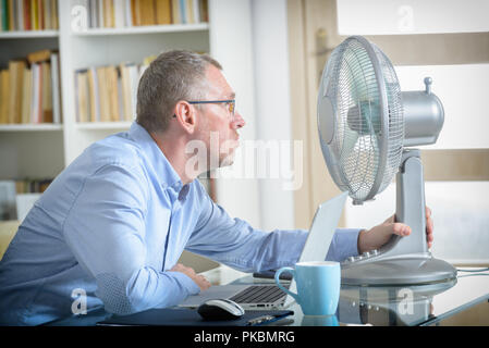 L'homme souffre de la chaleur tout en travaillant dans le bureau et tente de se rafraîchir par le ventilateur Banque D'Images