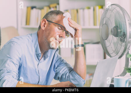 L'homme souffre de la chaleur tout en travaillant dans le bureau et tente de se rafraîchir par le ventilateur Banque D'Images