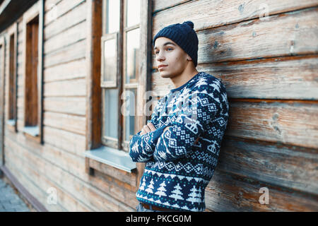 Beau jeune homme dans un pullover en tricot blanc et le chapeau à proximité d'une maison en bois. Banque D'Images