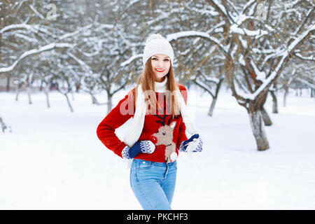 Belle fille élégante dans un chapeau en tricot, pull rouge avec un chevreuil et sur un fond blanc avec des arbres enneigés. Banque D'Images