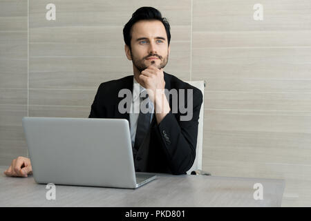 Portrait de beau jeune homme d'affaires assis au bureau, le travail avec ordinateur portable et à réfléchir sur son projet d'entreprise.. Banque D'Images