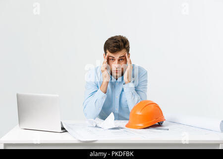 Portrait de l'ingénieur stress tenait la tête avec la main assis à la table et se sentir mal de tête au bureau sur fond isolé. Concept d'ingénierie. Banque D'Images
