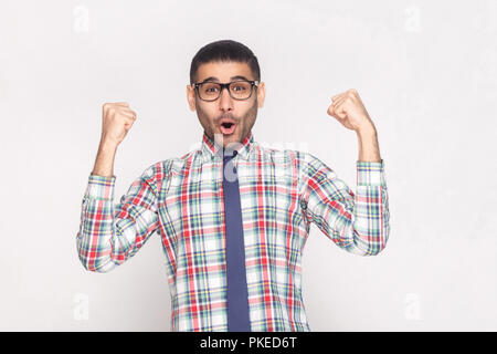 Heureux gagnant beau homme barbu en chemise à carreaux, cravate bleue et lunettes noires en criant et permanent à la caméra à main levée avec. Banque D'Images