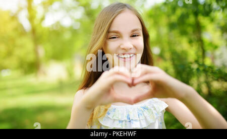 Cute preteen girl rire et tenant ses mains en forme de coeur sur lumineuse et ensoleillée journée d'été. Cute child s'amusant à l'extérieur. Heureux childhoo Banque D'Images
