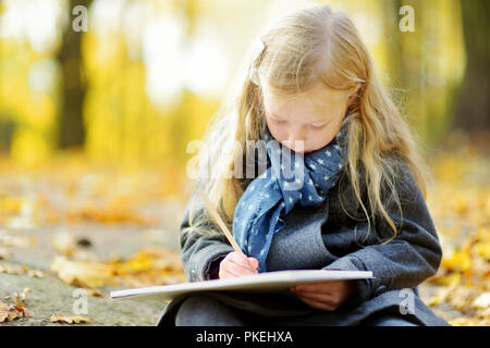 Cute little girl des croquis à l'extérieur sur la belle journée d'automne. Heureux l'enfant jouant dans le parc de l'automne. Dessin pour enfants avec des crayons de couleur. Activités d'automne Banque D'Images