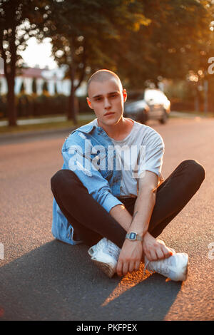 Jeune homme élégant brutal avec vêtements en jean dans un T-shirt gris, pantalon noir, chaussures blanc se trouve sur une route asphaltée au coucher du soleil Banque D'Images