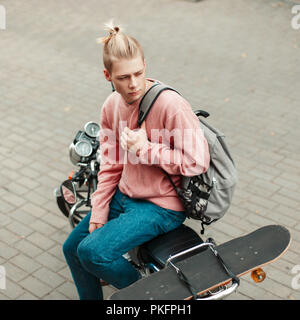 Beau jeune homme avec une coiffure dans un pull rose avec un sac à dos et d'un skateboard assis sur une moto Banque D'Images