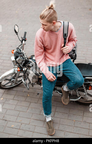 Beau jeune homme dans un chandail de mode rose et bleu jeans avec un sac à dos est assis sur une moto Banque D'Images