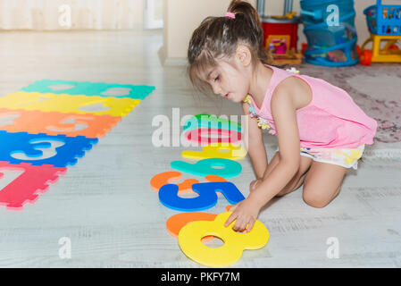 Vue latérale d'une mignonne petite fille est assise et tapis de jeu de puzzle coloré tient avec des nombres à la maison. Banque D'Images