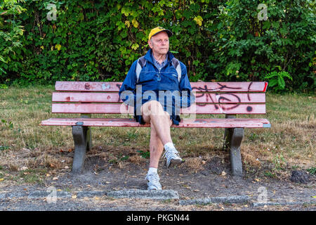 Berlin,Dahlem, Senior man sitting on couverts de graffitis vieux banc l'air inquiet. Un homme âgé portant des shorts & cap est assis sur un banc Banque D'Images