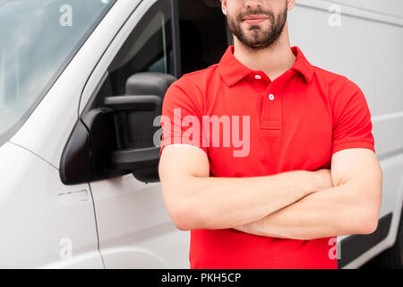 Cropped shot of delivery man en uniforme rouge avec les bras croisés debout à van Banque D'Images