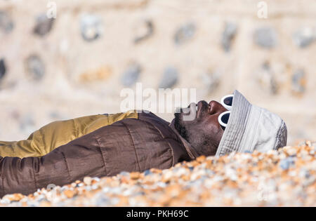 Homme noir portant sur une plage sur une chaude journée ensoleillée au printemps portant un chapeau, manteau et lunettes de soleil, au Royaume-Uni. Banque D'Images