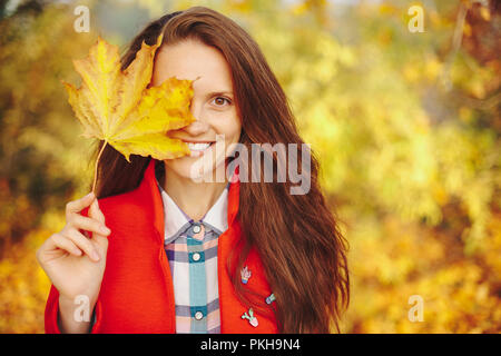 Belle jeune femme à la longue chevelure ondulée couvrant le visage avec une feuille en souriant contre un fond jaune d'or. La peau douce et sourire en santé. Re Banque D'Images