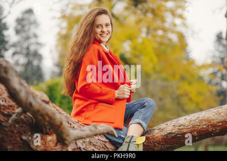 Belle jeune femme à la longue chevelure ondulée en veste rouge et jeans assis sur une branche d'arbre. Smiling girl portrait d'automne. Banque D'Images