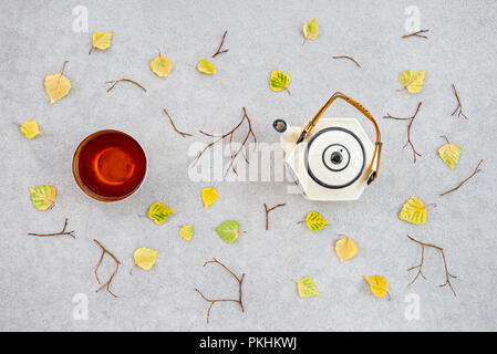 Thé chaud lors d'une fraîche journée d'automne. Tasse, théière et feuilles tombées sur fond de béton. Banque D'Images