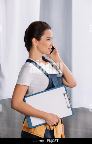 Vue de côté de belles jeunes repairwoman avec presse-papiers de parler par téléphone Banque D'Images