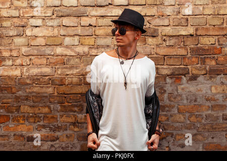 Bel homme élégant dans un t-shirt blanc, veste en cuir noire et un chapeau avec des lunettes qui pose près de old brick wall Banque D'Images
