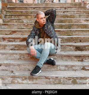 Bel homme élégant dans une veste militaire avec un sweat et un jean est assis sur les marches Banque D'Images