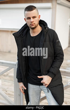 Modèles beau jeune homme dans un manteau d'hiver noir à la mode se faisant passer l'extérieur Banque D'Images