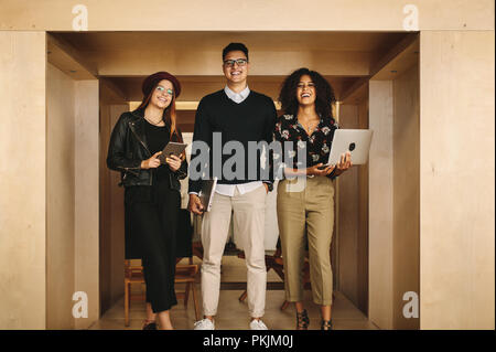 Heureux coéquipiers standing in office holding des ordinateurs portatifs et d'un comprimé. Businessman avec deux collègues femmes au travail debout tenant un ordinateur portable. Banque D'Images