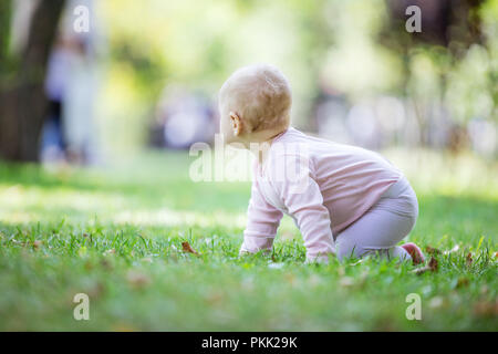 Petite fille blonde rampent sur pelouse au parc et à la recherche de quelque chose de côté Banque D'Images