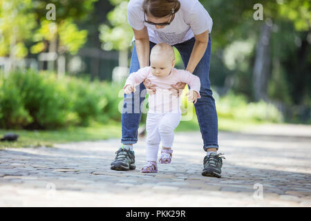 Le soutien mère fille de bébé et l'aider à faire vos premiers pas. Mère et fille sur marche en parc d'été. Banque D'Images