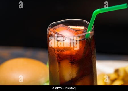 Close up sur verre de soude avec de la glace de petits cubes de glace et de paille vert coincé en elle alors que burger et des frites s'asseoir en arrière-plan Banque D'Images