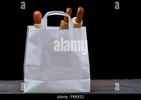 Vue latérale du petit sac blanc contenant des hot dogs enveloppés dans du pain assis sur table en bois en face de fond noir Banque D'Images