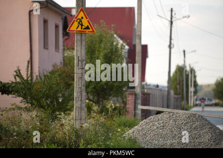 Signalisation triangulaire jaune vif  ; attention, travaux à venir, posté en tête de pilier en béton dans l'herbe verte au tas de gravier sur le paysage rural, suburba Banque D'Images