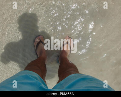 Les jambes avec un short bleu et de porter des tongs, debout dans l'eau claire de la mer en Thaïlande. Banque D'Images