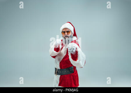 Funny guy boxe agressive avec noël hat posing at studio. Maison de vacances de Nouvel An. Noël, x-mas, hiver, cadeaux concept. Homme portant costume père noël sur gris. Copier l'espace. Les soldes d'hiver. Banque D'Images