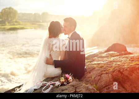 Belle amour couple kissing, assis sur des rochers près de la rivière. Couple de mariés au coucher du soleil et d'eau, l'amour et de tendres sentiments. Couple aimant se reposer. Banque D'Images