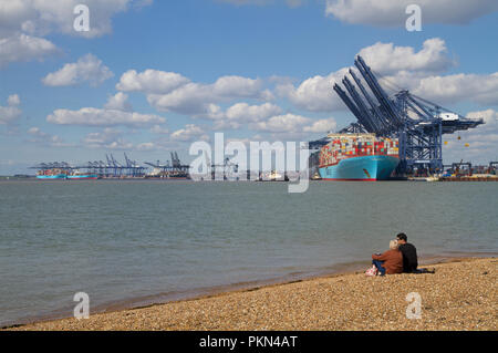 Couple assis sur la plage de galets à Felixstowe donnant sur l'eau à l'grandes grues à conteneurs et navires ont accosté au port. Banque D'Images