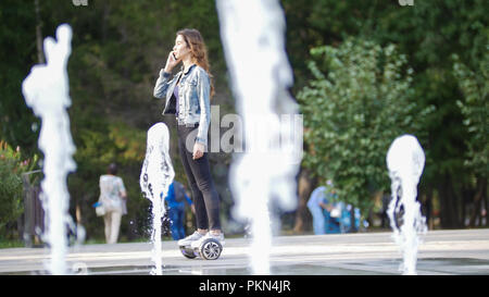 Jeune fille monte un gyro et parle au téléphone dans le parc un jour d'été, dans l'avant-plan des Fontaines Banque D'Images