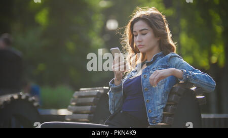 Jeune jolie jeune fille assise sur un banc, de tenir le téléphone, une jambe tenue sur un gyro Banque D'Images