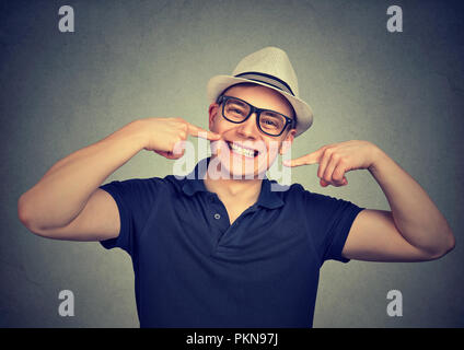 Jeune homme élégant avec chapeau et lunettes en montrant des dents blanches lumineuses donnant sourire à huis clos sur fond gris Banque D'Images