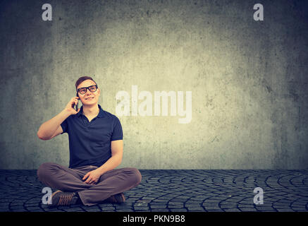 Casual young man sitting sur fond gris et se faire appeler sur smartphone smiling up Banque D'Images