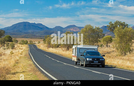 Voiture avec caravane sur route menant à travers des paysages boisés et les herbes d'or à des pointes de Warrumbungle National Park s'élevant dans le ciel bleu dans le NSW Banque D'Images