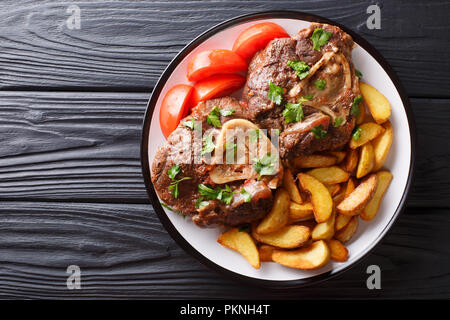 Ossobuco jarret avec une garniture de tranches de pommes de terre frites et les tomates sur une assiette sur la table. Haut horizontale Vue de dessus Banque D'Images