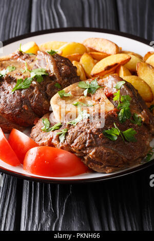 Menu italien : steak de veau braisé Ossobuco avec pommes de terre sautées et des tomates sur une plaque verticale sur la table. Banque D'Images