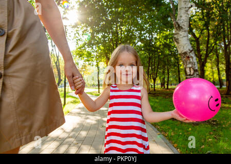 Close-up shoot de jolie petite blonde aux cheveux long enfant fille en robe colorée tenant la main en toute confiance de jeune mère, et rose dans l'autre main à l'extérieur, sur fond d'été ensoleillé coloré brouillée. Banque D'Images