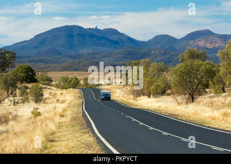 Voiture avec caravane sur route menant à travers des paysages boisés et les herbes d'or à des pointes de Warrumbungle National Park s'élevant dans le ciel bleu dans le NSW Banque D'Images