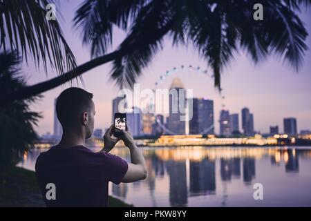 Jeune homme (touristique) prise de photo de la ville par téléphone mobile. Paysage urbain de Singapour au coucher du soleil. Banque D'Images