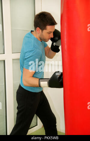 Male boxer de la formation est l'entraînement avec sac de boxe au gymnase club. Portrait d'une jeune athlète beau homme sports boxe gants rouge dans la pratique Banque D'Images