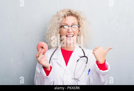 Jeune blonde dietist médecin femme de plus grunge grey wall eating pointant et montrant avec le pouce jusqu'à la côte avec happy face smiling Banque D'Images