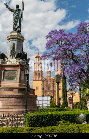 Statue de l'indépendance et le chef Miguel Hidalgo Parroquia Nuestra Señora de Dolores Eglise Catholique dans la Plaza Principal avec la floraison des jacarandas derrière en Dolores Hidalgo, Guanajuato, Mexique. Hildago était un prêtre de paroisse qui a publié le désormais célèbre Grito - un appel aux armes pour l'indépendance de l'Espagne. Banque D'Images