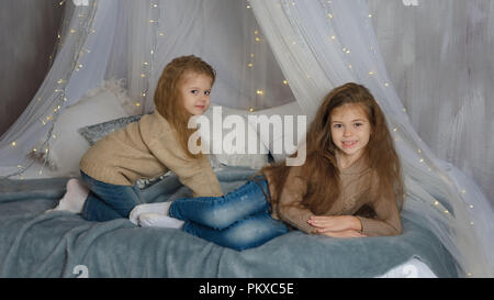 Deux petites soeurs sont en attente de Noël. Les filles s'allonger sur le lit avec une guirlande. Ils sont en jeans et pulls. Enfance heureuse. Banque D'Images